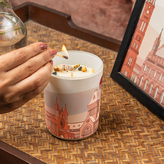 Hand lighting a candle in a decorative container with a building design on a woven surface.